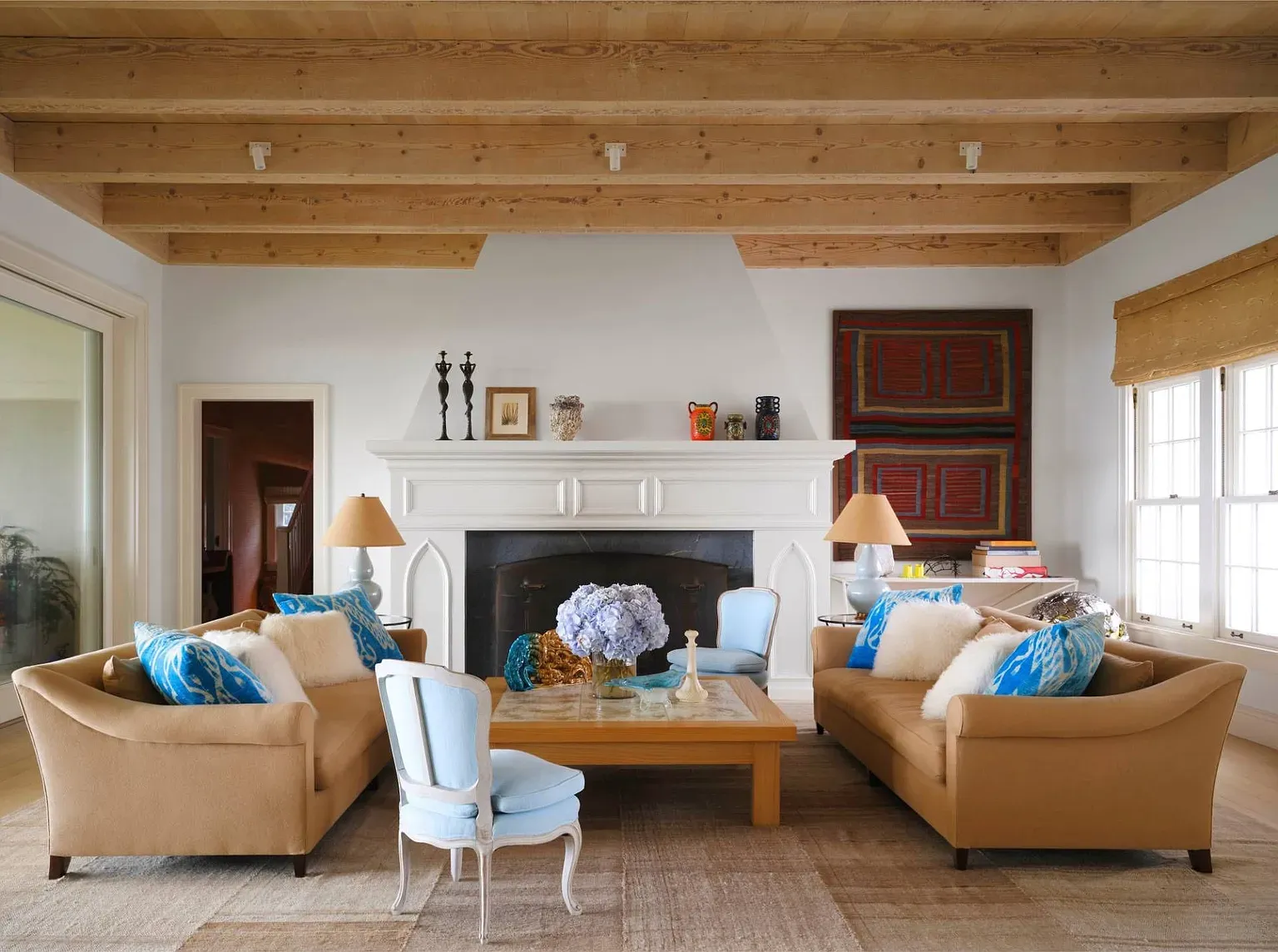 "Here’s the living room! The wooden beams overhead really add some warmth, and that fireplace is perfect for gathering around. I love how the blue accents pop against the soft tones of the furniture. It feels so inviting!"