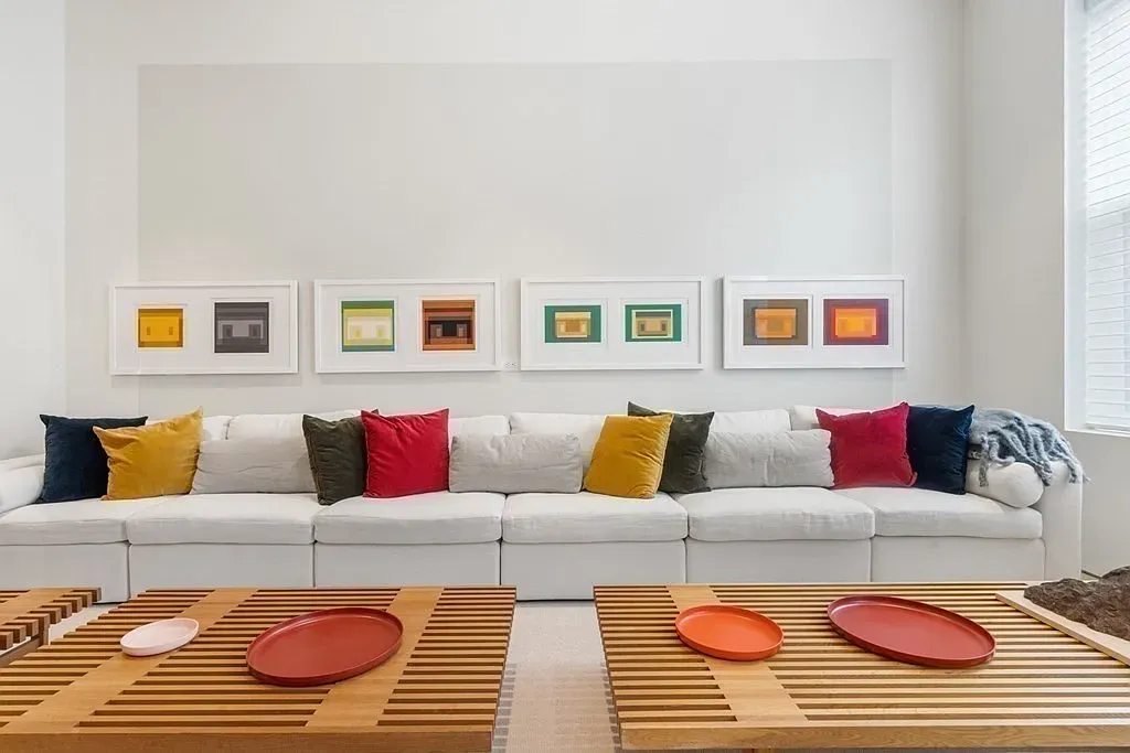 "Take a look at this comfy living area! I love how the colorful pillows pop against the white sofa, and those framed prints bring such a fun energy to the space. The coffee tables are really unique too; they add a nice touch of warmth